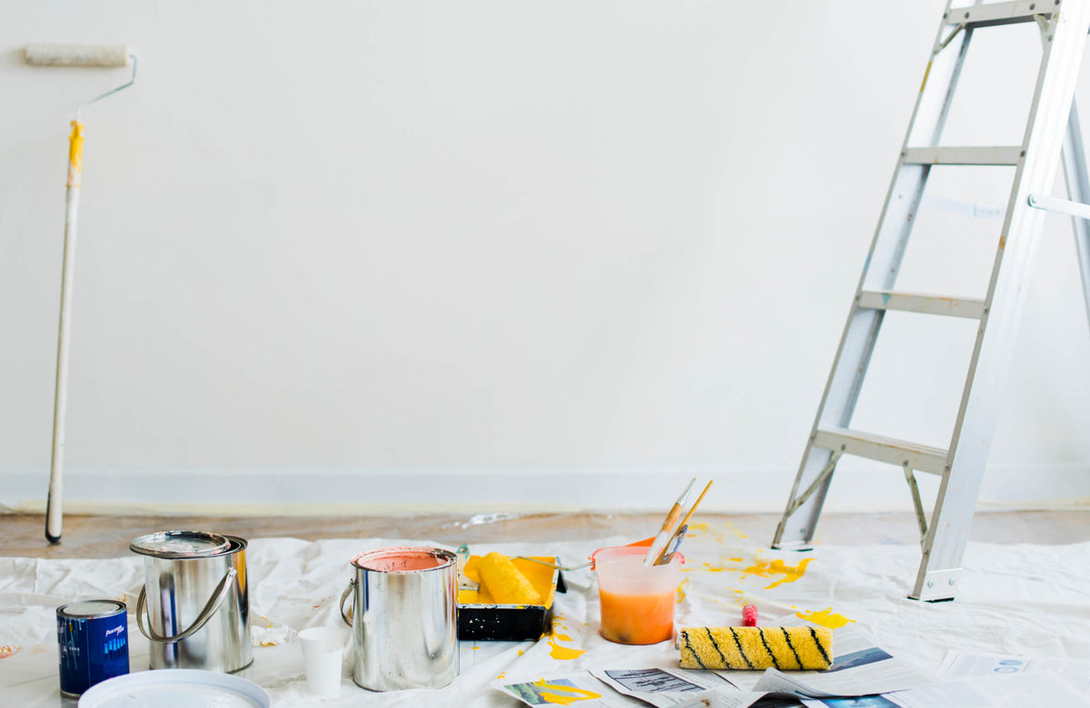 A diy bathroom remodel scene in progress shows a freshly painted white wall with a ladder nearby, surrounded by paint cans, rollers, brushes, and splashes of yellow paint laid out on protective drop cloths.
