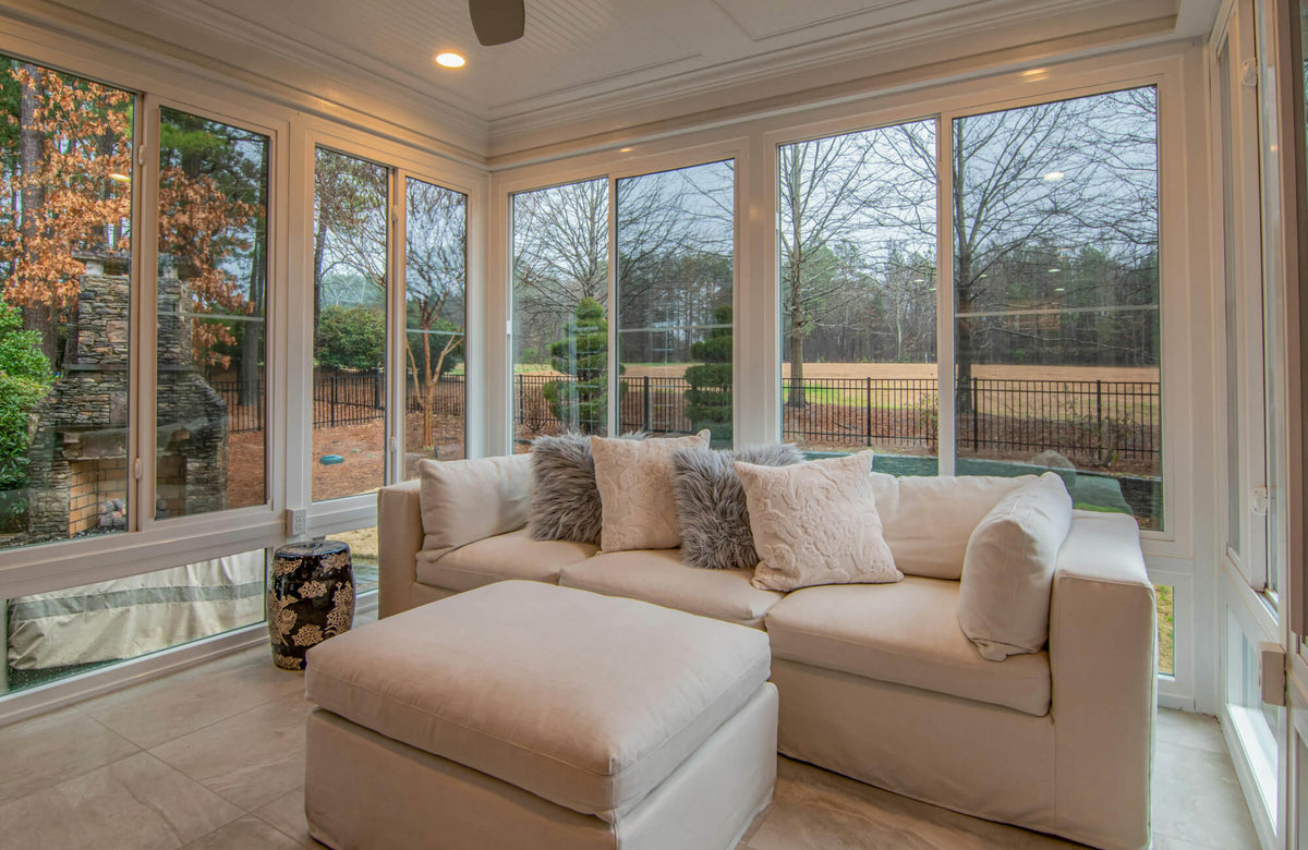 Contemporary sunroom with plush sectional sofa, decorative pillows, large windows, and light stone tile flooring overlooking a serene outdoor landscape.