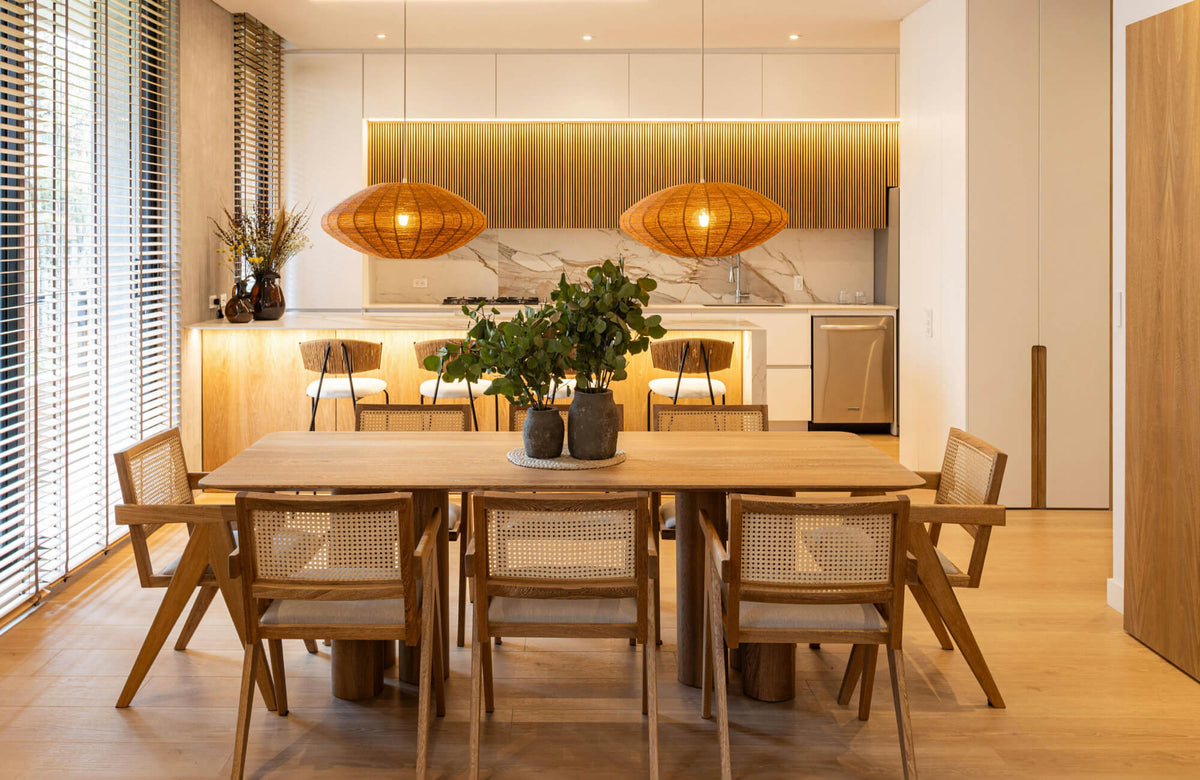Warm, modern interior showcasing a minimalist dining room table set with a solid wood rectangular table and woven-back chairs, centered beneath sculptural pendant lights and seamlessly connected to a light-filled kitchen.