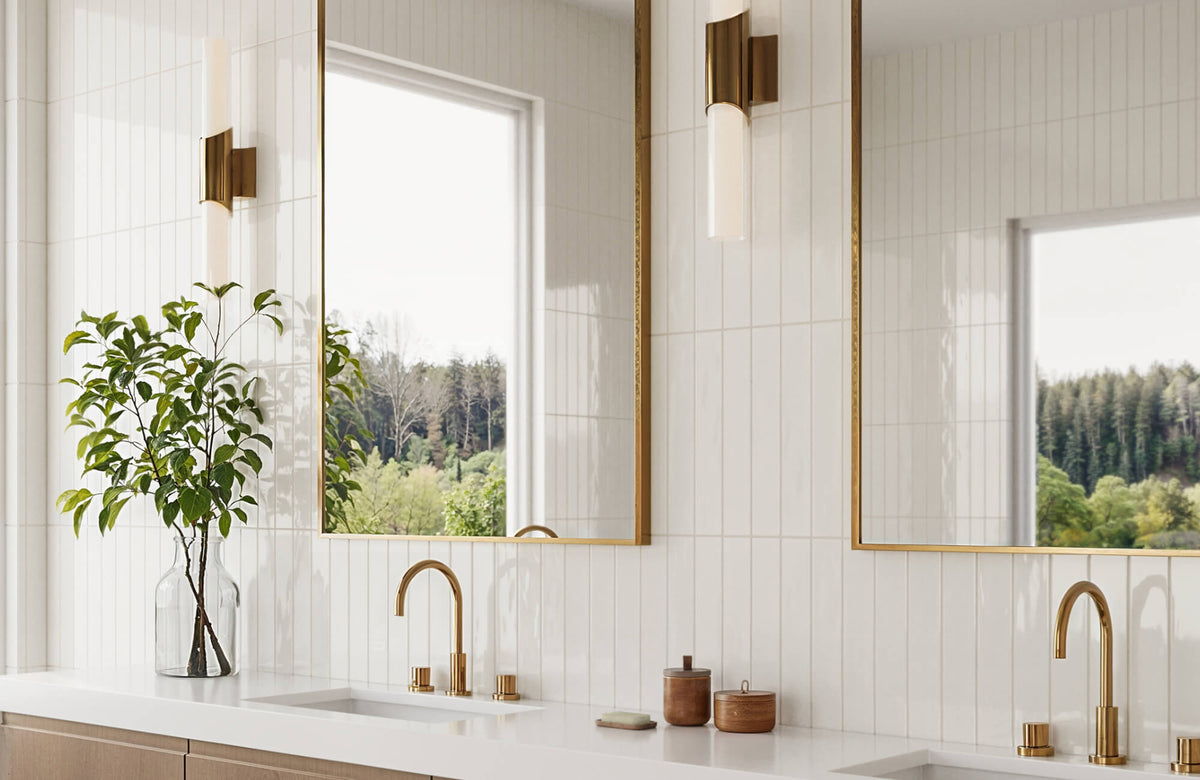 Bright bathroom vanity with glossy white vertical subway tiles, brass mirrors and fixtures, and a glass vase of greenery beside a long white countertop.