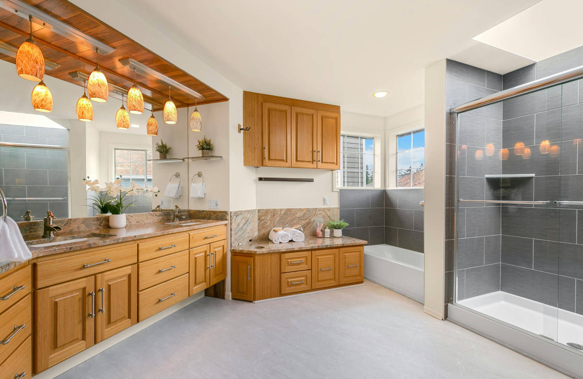 Spacious modern bathroom with oak vanity, granite countertop, frameless glass shower, gray tile walls, and warm pendant lighting.