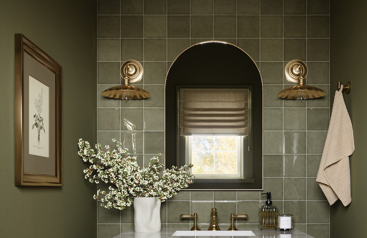 Elegant bathroom with arched mirror above sink, framed by gold sconces against green tiled wall.