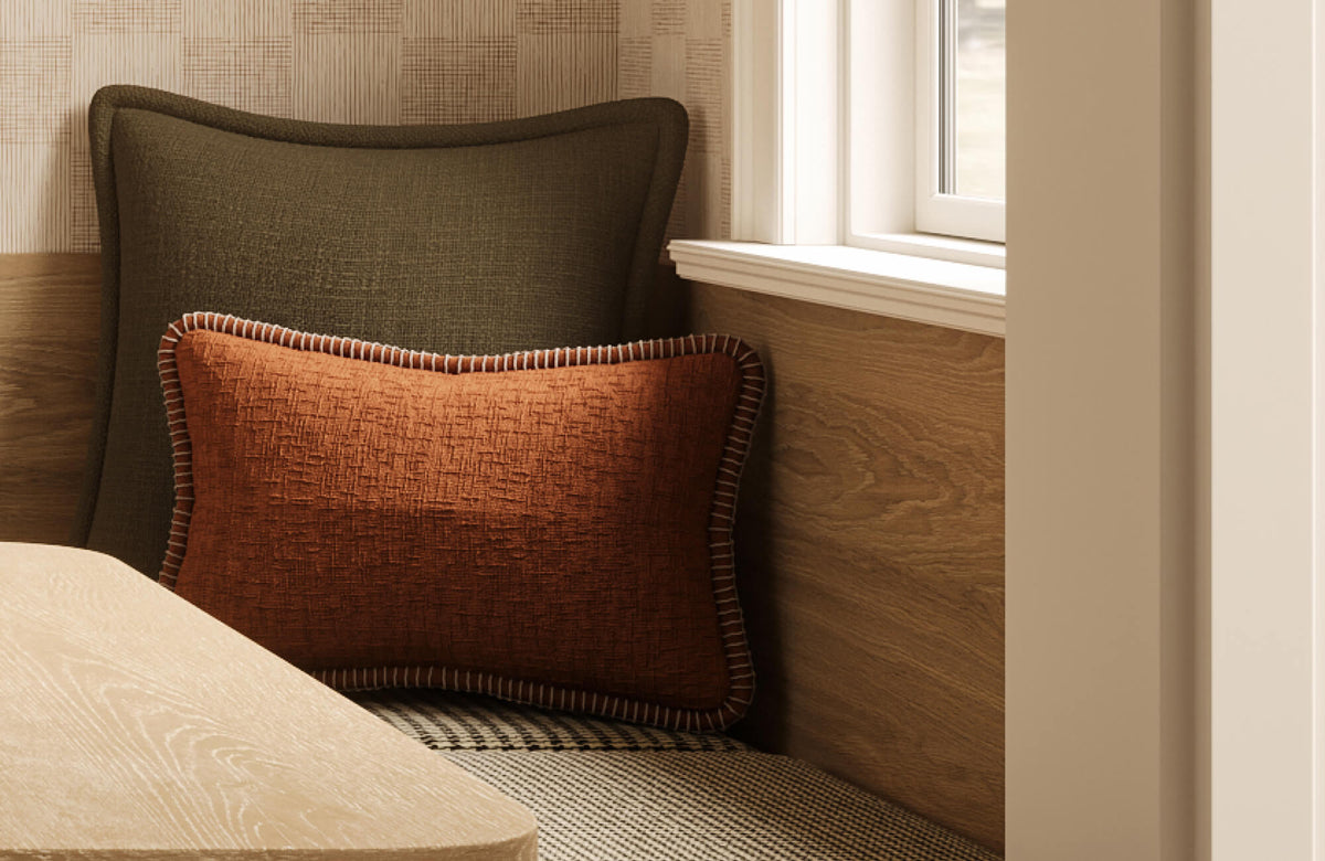 Rust-colored lumbar throw pillow with contrast whipstitch trim layered against a dark olive accent pillow on a built-in bench, creating a warm and structured seating arrangement beside a sunlit window.