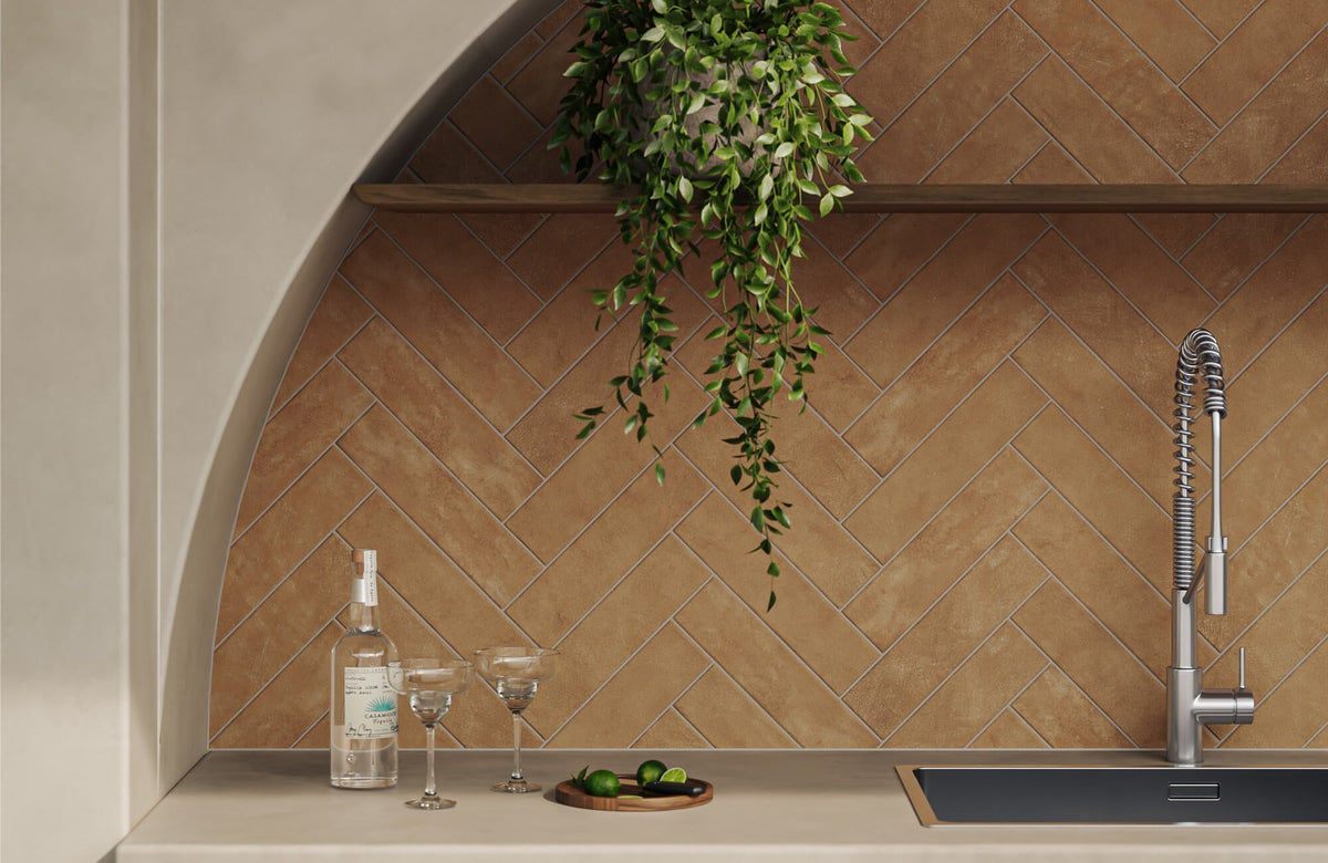 Warm brown herringbone tile backsplash in an arched outdoor bar area with a stone countertop, chrome faucet, wood shelf, trailing greenery, and glassware.