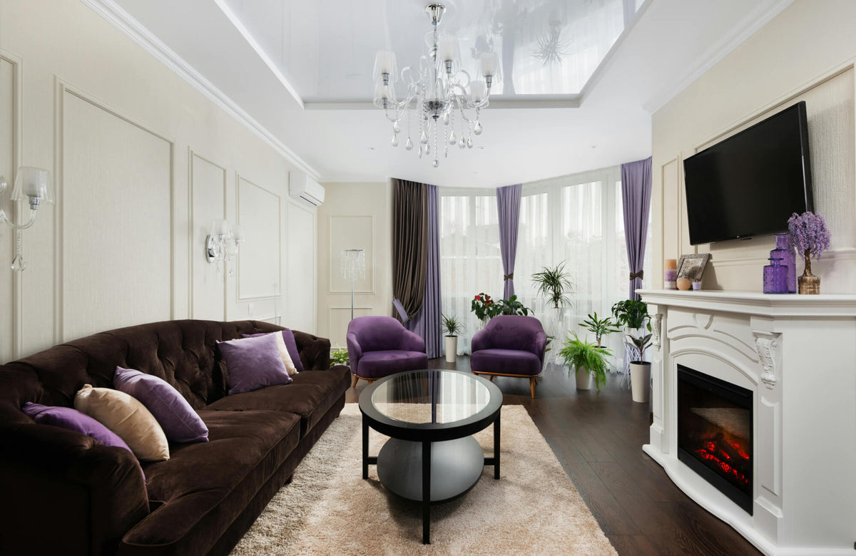 Elegant living room with dark wood floors, white fireplace, crystal chandelier, and plush purple seating with decorative pillows.