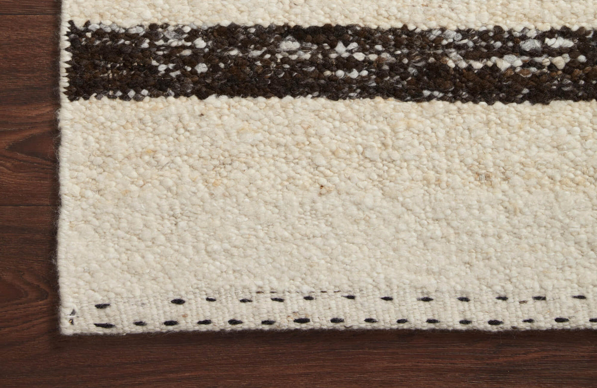 Close-up of a cream textured wool rug with a dark horizontal stripe, stitched border detail, and rich wood flooring visible along the edge.
