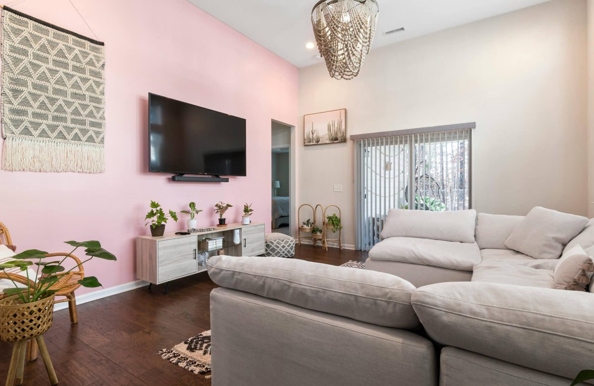 Bright living room with blush pink accent wall, woven textile art, wall-mounted TV, neutral sofa, plants, and modern chandelier.