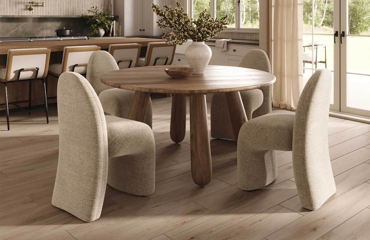 Warm dining area and kitchen with wood-looking floor tile, a round wooden table, upholstered chairs, cream curtains, and a softly lit kitchen island in the background.