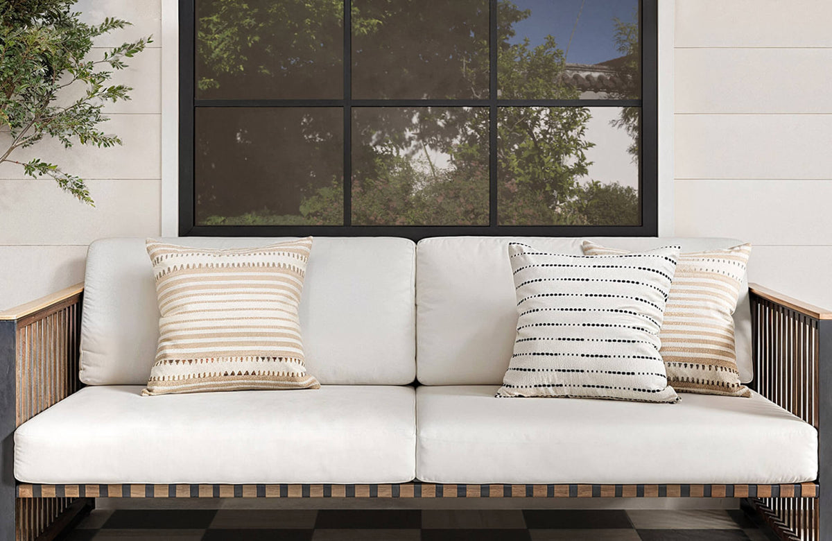 Outdoor sofa with white cushions styled with three square throw pillows in beige, cream, and black striped patterns beneath a large black-framed window.