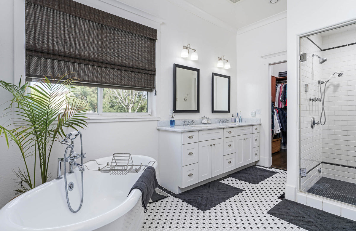 Bright white bathroom with clawfoot tub, dual vanity, black-framed mirrors, mosaic tile floor, woven blind, and glass shower enclosure.