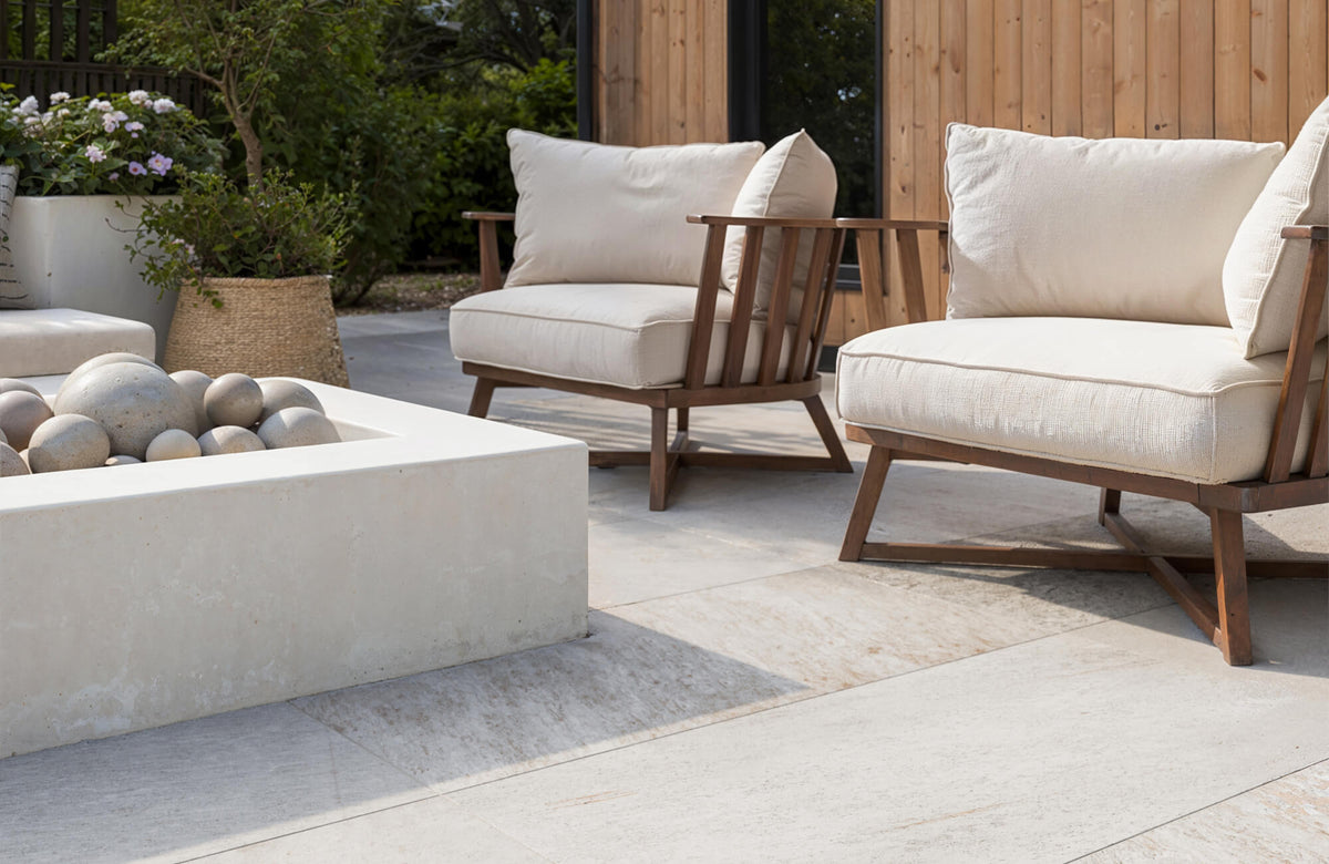 Sunlit outdoor living space featuring pale stone-inspired tiles underfoot, paired with wood-framed lounge chairs, soft neutral cushions, and surrounding greenery.