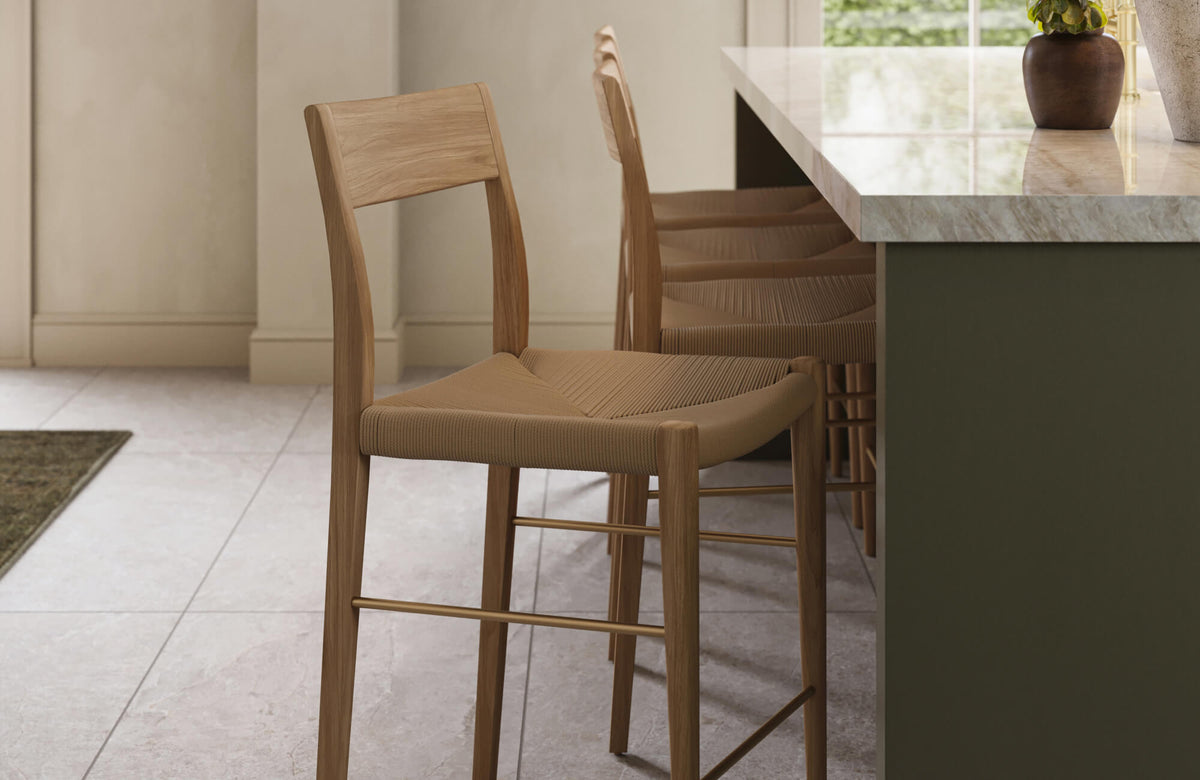 Oak counter stools with softly curved wooden frames and woven tan seats tucked under a green kitchen island, paired with a light stone countertop and neutral tiled flooring.