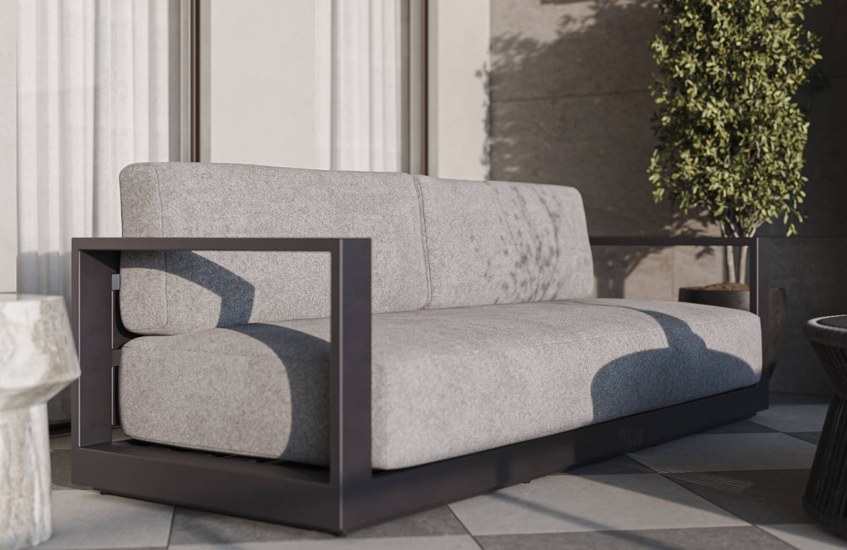 Contemporary outdoor patio featuring a modern gray cushioned sofa with a black geometric metal frame, checkerboard tile flooring, sculptural side tables, and surrounding greenery in a minimalist open-air setting.