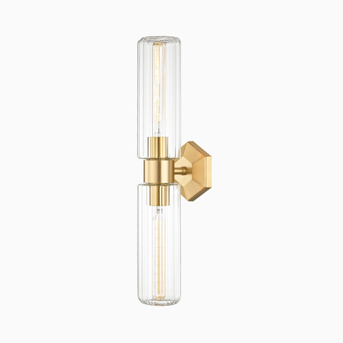 Wall sconce with aged brass finish, hexagonal backplate, and twin vertically fluted glass columns for soft, ambient glow.