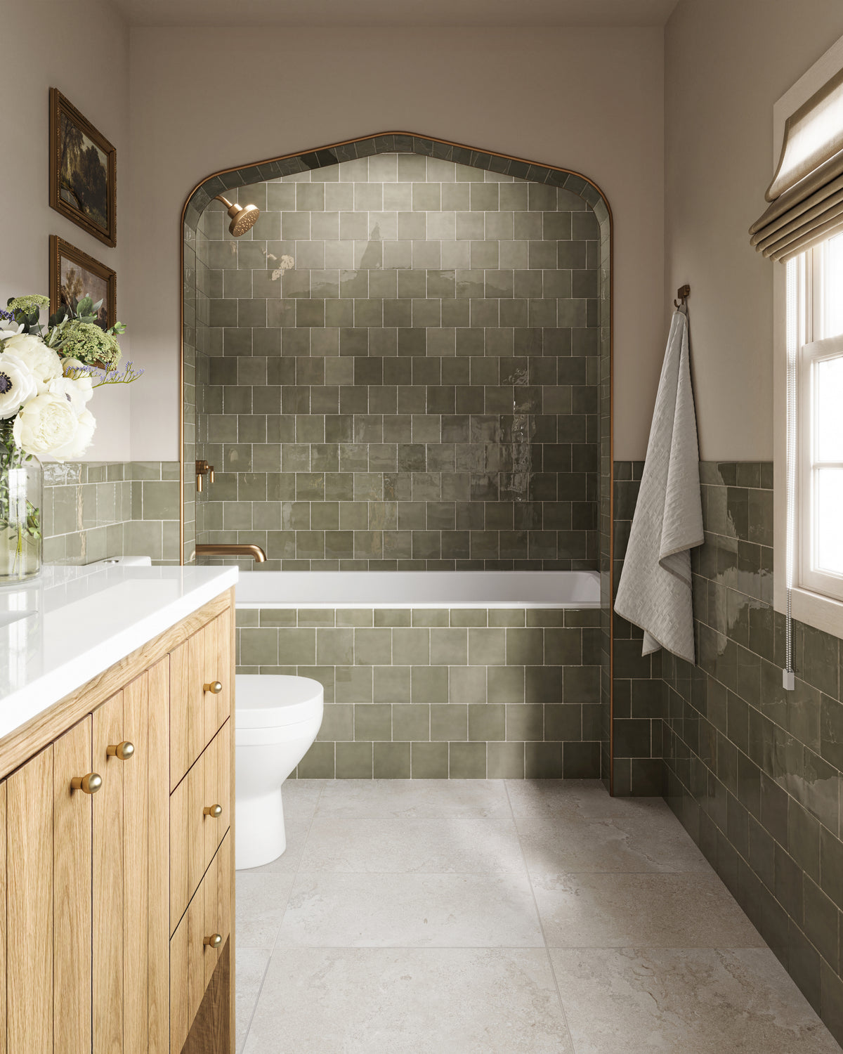 Bathroom with light natural oak vanity and white quartz countertop beside an arched bathtub alcove lined with glossy olive green subway tiles, brass shower fixtures, a wall-mounted towel, and a sunlit window.