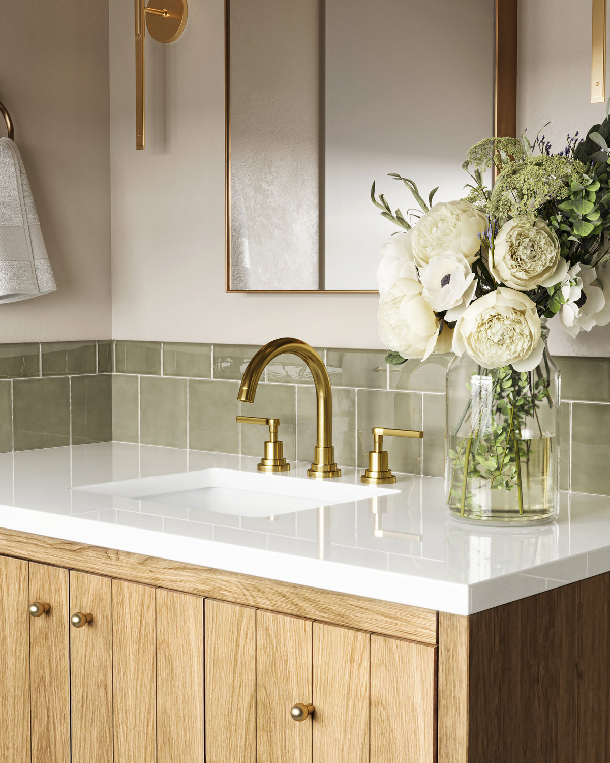 Close-up of a light natural oak bathroom vanity with vertical wood grain cabinet doors and brass knobs, topped with a white quartz countertop, integrated sink, and brushed brass faucet. A glass vase filled with white flowers sits beside the sink against glossy sage green subway tile and a brass-framed mirror above.