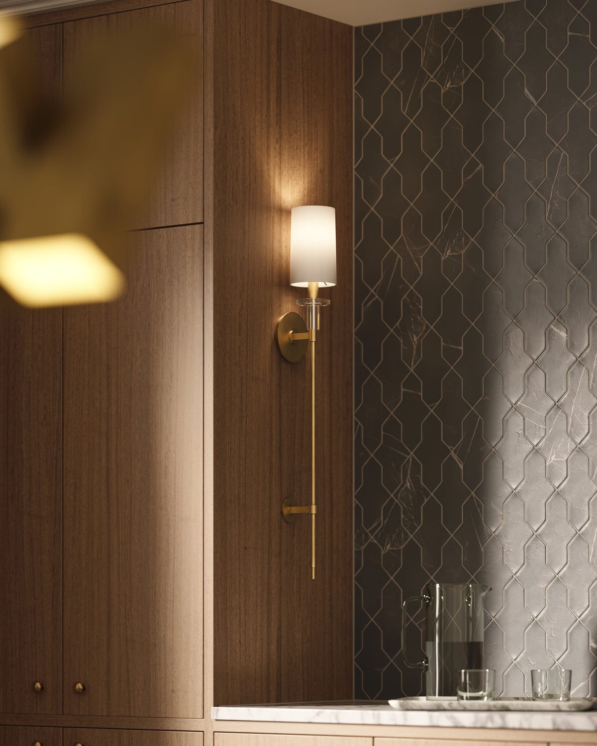 Aged brass wall sconce with white shade mounted on warm wood panel wall beside dark geometric tile backsplash.