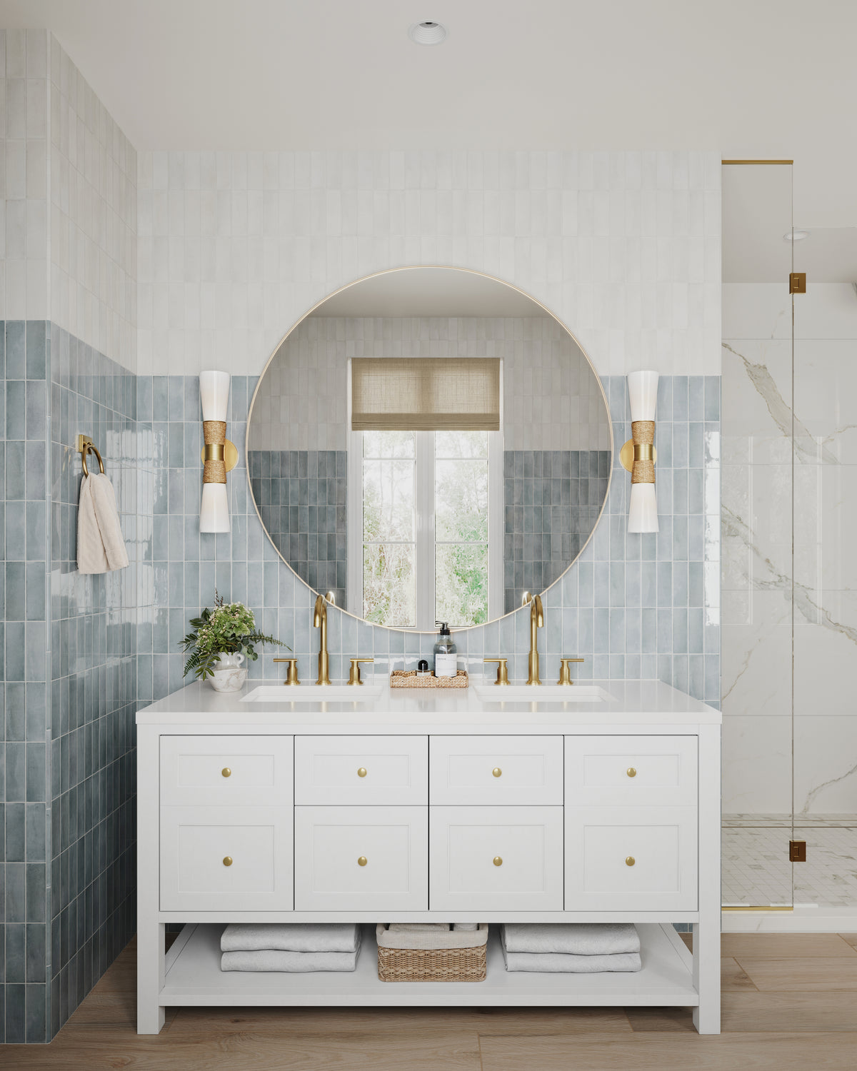 White double bathroom vanity with a white quartz countertop, gold faucets, and brass hardware, featuring multiple drawers and an open lower shelf with folded towels and a basket. A large round mirror with gold wall sconces sits above blue-gray tile wainscoting, with a glass shower enclosure beside the vanity.