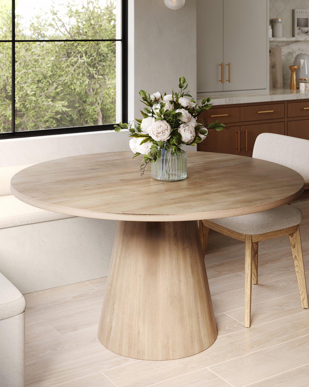 Round light oak dining table with pedestal base styled with white florals, paired with upholstered chairs in a bright, modern kitchen nook.