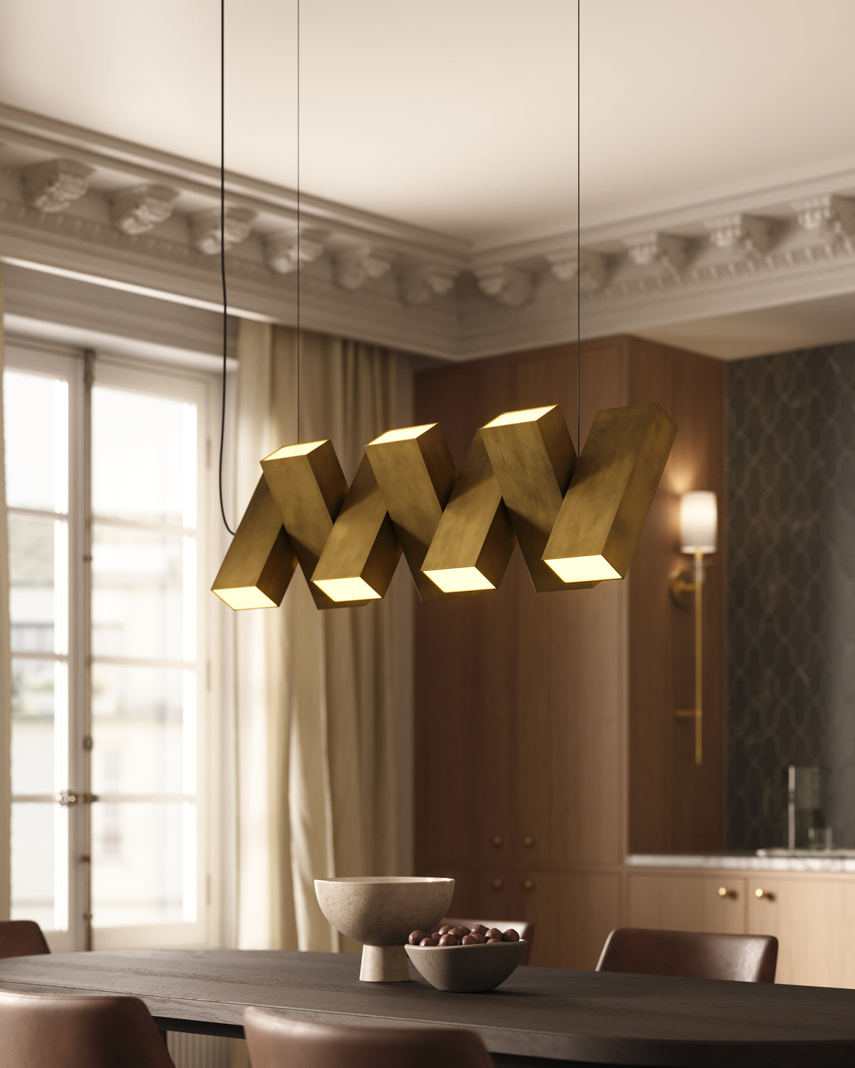 Elegant dining room featuring a vintage brass geometric pendant light with sculptural rectangular forms and warm integrated LED glow, suspended above a dark wood table with leather chairs, set against classic crown molding and rich wood paneling.