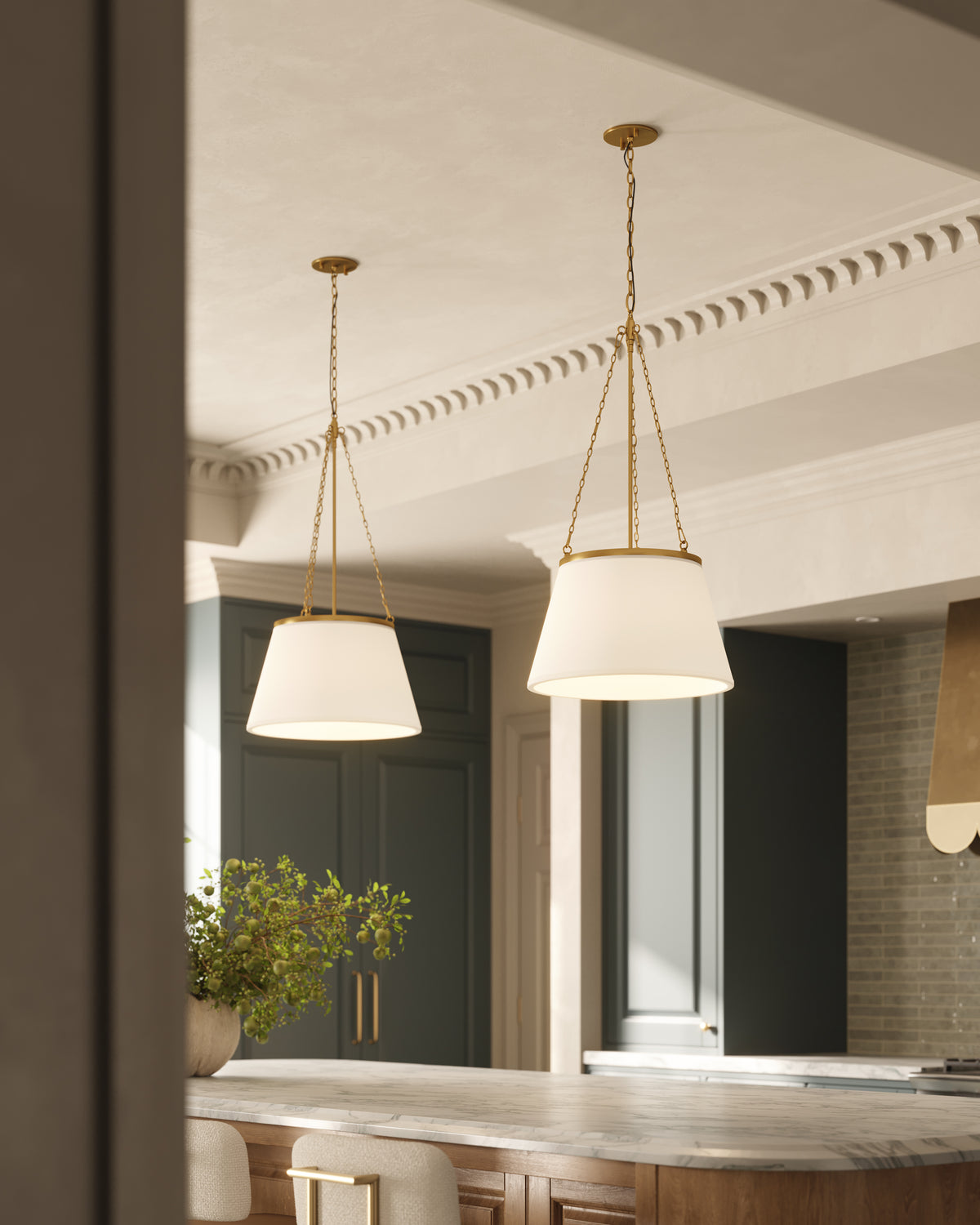 Brushed gold chain hung pendant lights with white fabric shades suspended above a marble kitchen island, casting soft light across cabinetry and crown molding.