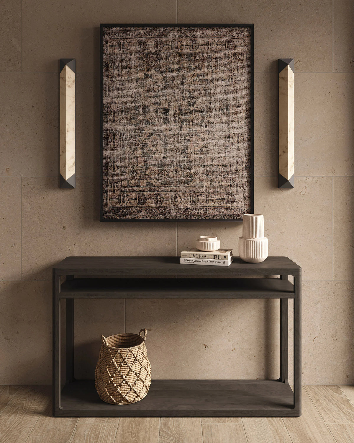 Wall art with a softly faded taupe and dark gray pattern hangs on a neutral stone wall, flanked by slim vertical wall sconces. Below, a dark console table holds stacked books, sculptural ceramic vases, and a woven basket on the lower shelf, set over light wood flooring for a calm, layered look.