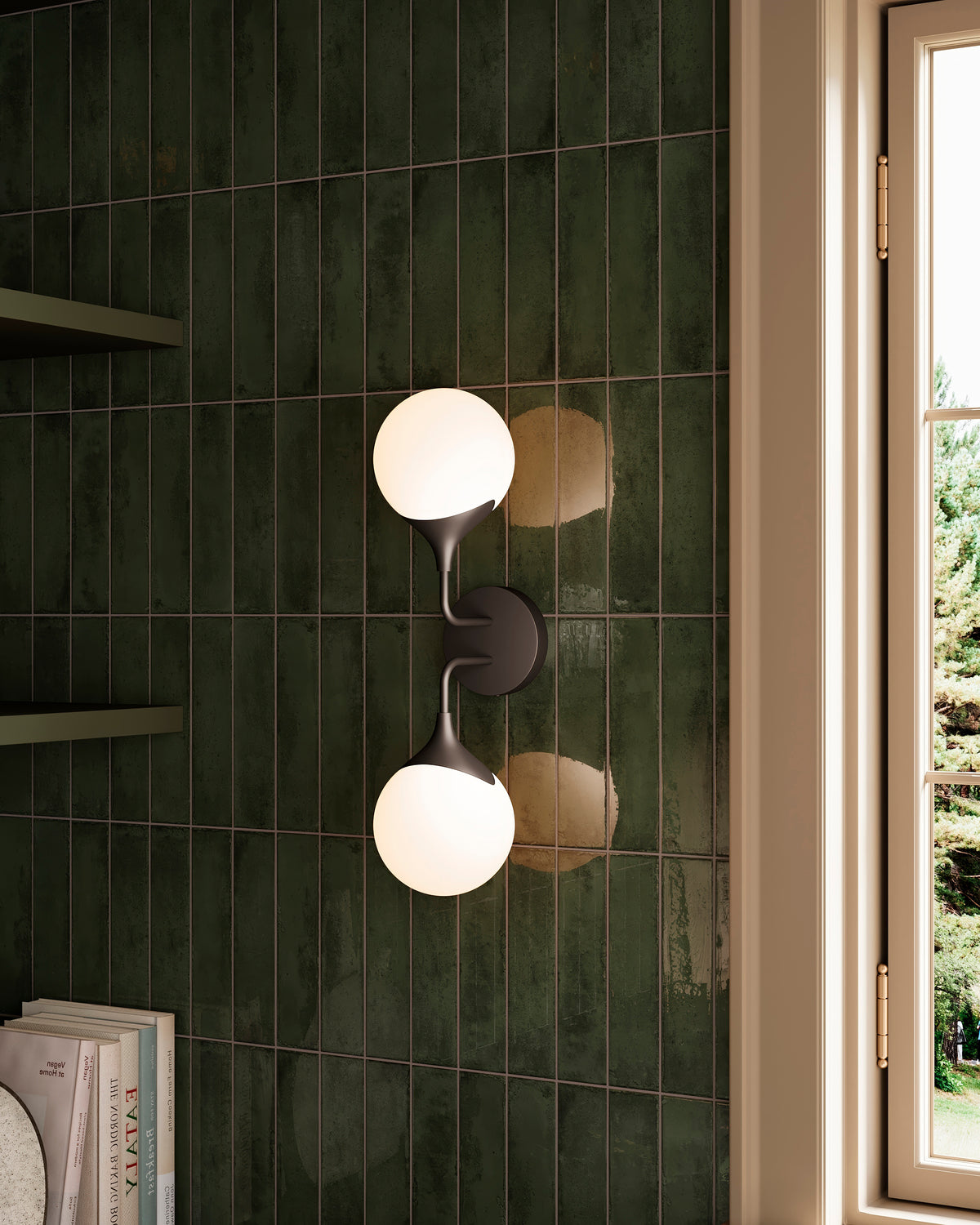 Matte black two light vanity wall fixture with frosted glass globe shades mounted on glossy green tile walls beside a window, casting soft ambient light.