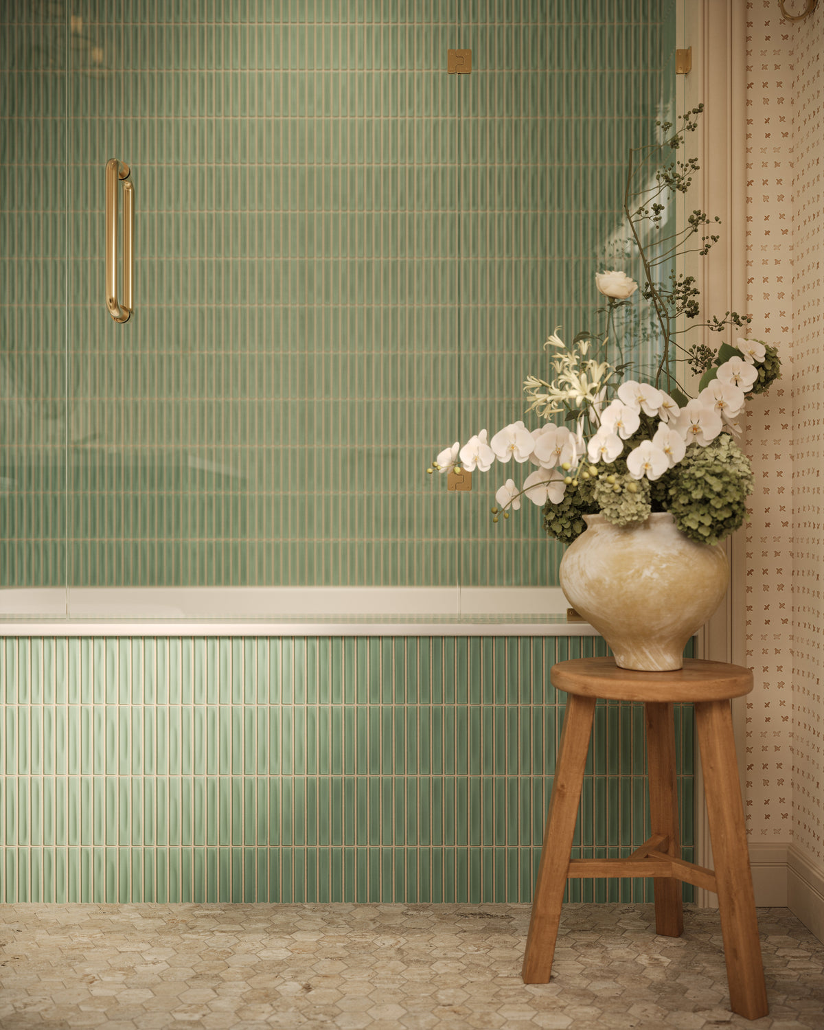 Bathroom vignette with sage green kitkat mosaic-look tile covering the shower wall and tub apron, paired with brass hardware, patterned wallpaper, stone mosaic flooring, and a wooden stool holding a ceramic vase of white flowers.
