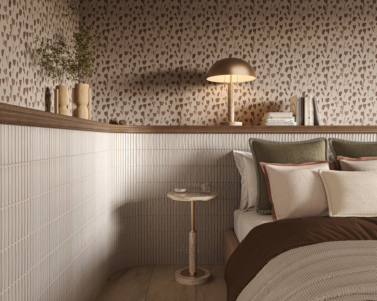 Cozy bedroom corner with crisp white glossy kitkat mosaic-look tile used as wall wainscoting, paired with patterned beige wallpaper, a wood picture ledge, layered neutral bedding, a small round bedside table, and a warm table lamp.