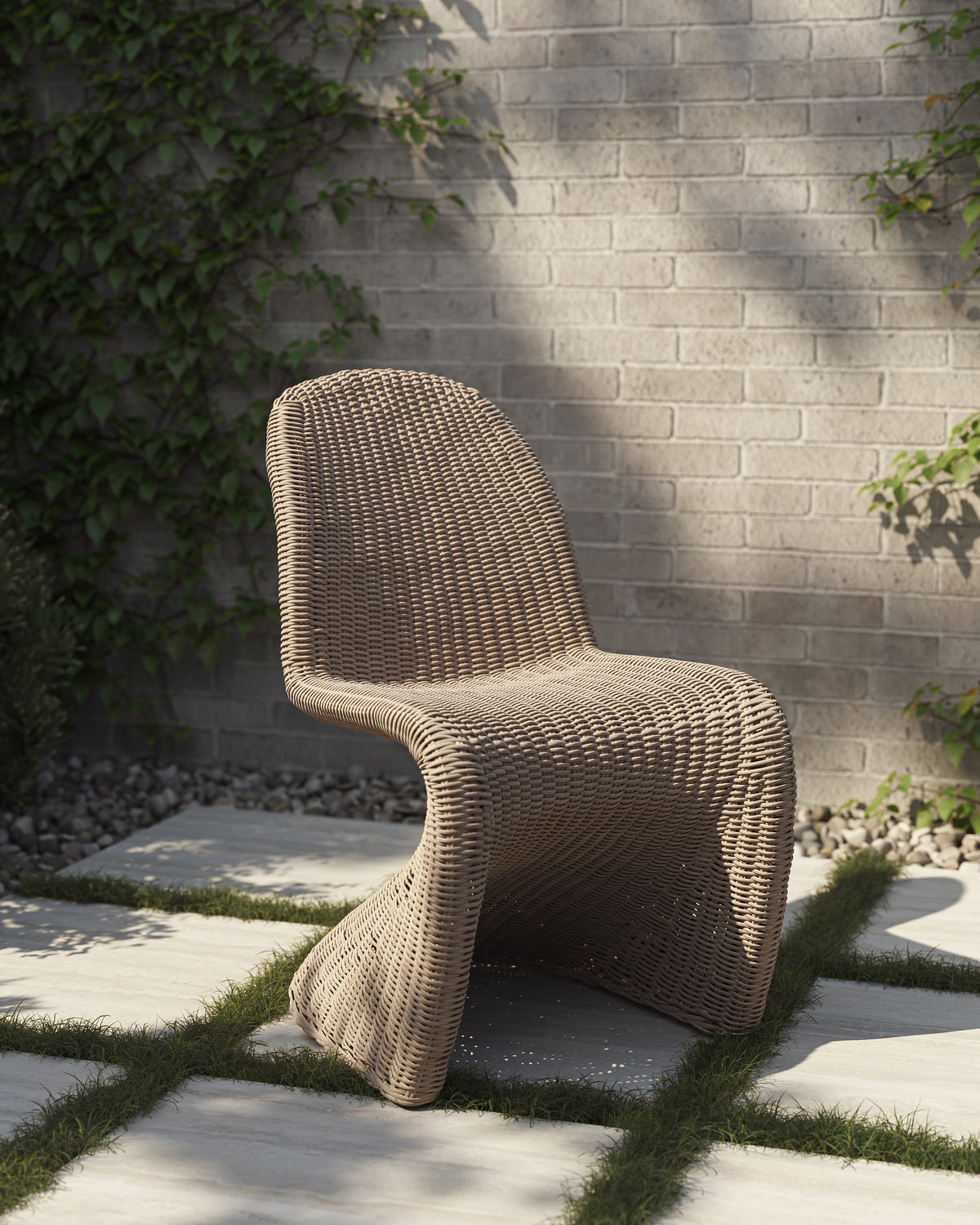 White wicker outdoor dining chair with a curved sled base and arched back set on pale stone pavers beside a gray brick wall, clipped lawn joints, and leafy garden plants.