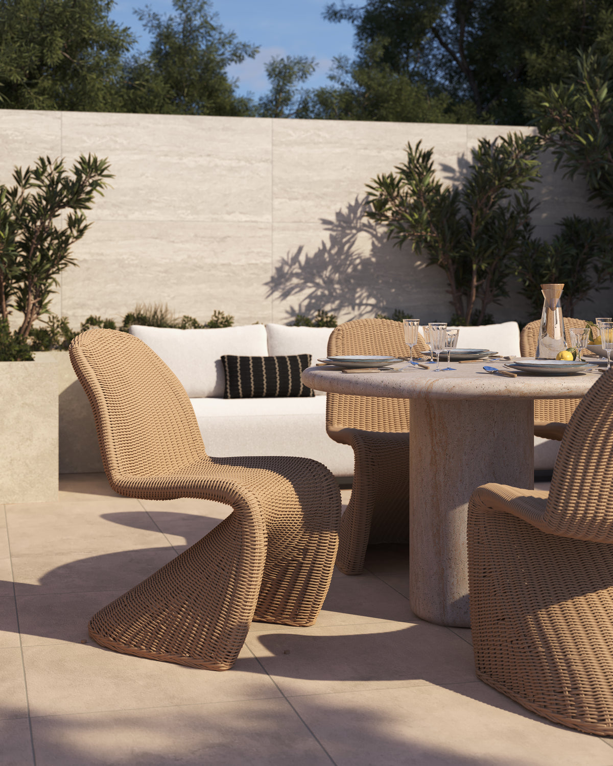 Natural wicker outdoor dining chair with a curved sled base and arched back styled beside a round beige stone dining table on a sunlit patio with pale floor tile and cream stone walls.