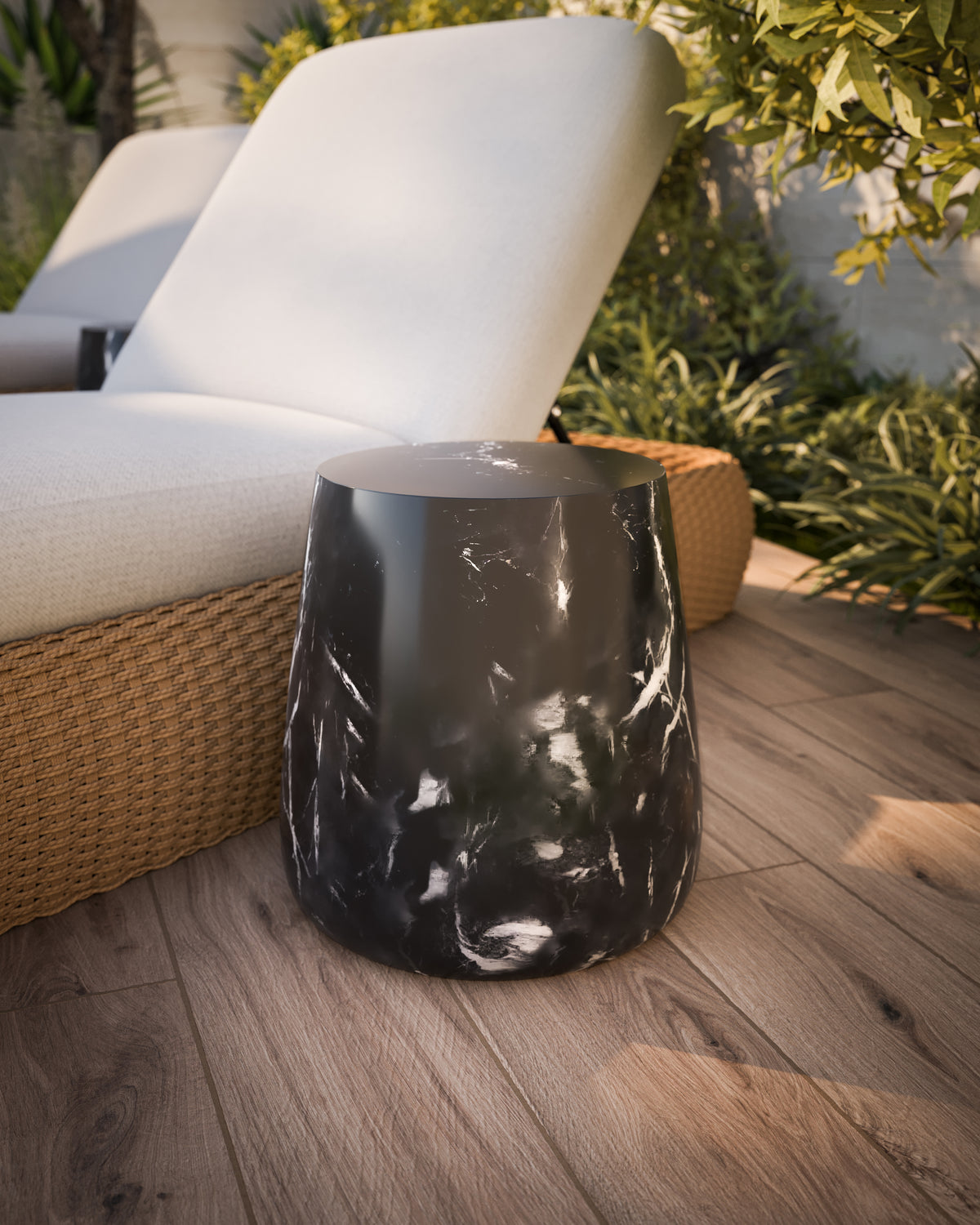 Our Dante Outdoor End Table in Black styled beside a cream cushioned wicker sun lounger on a warm wood plank deck surrounded by green garden plants.