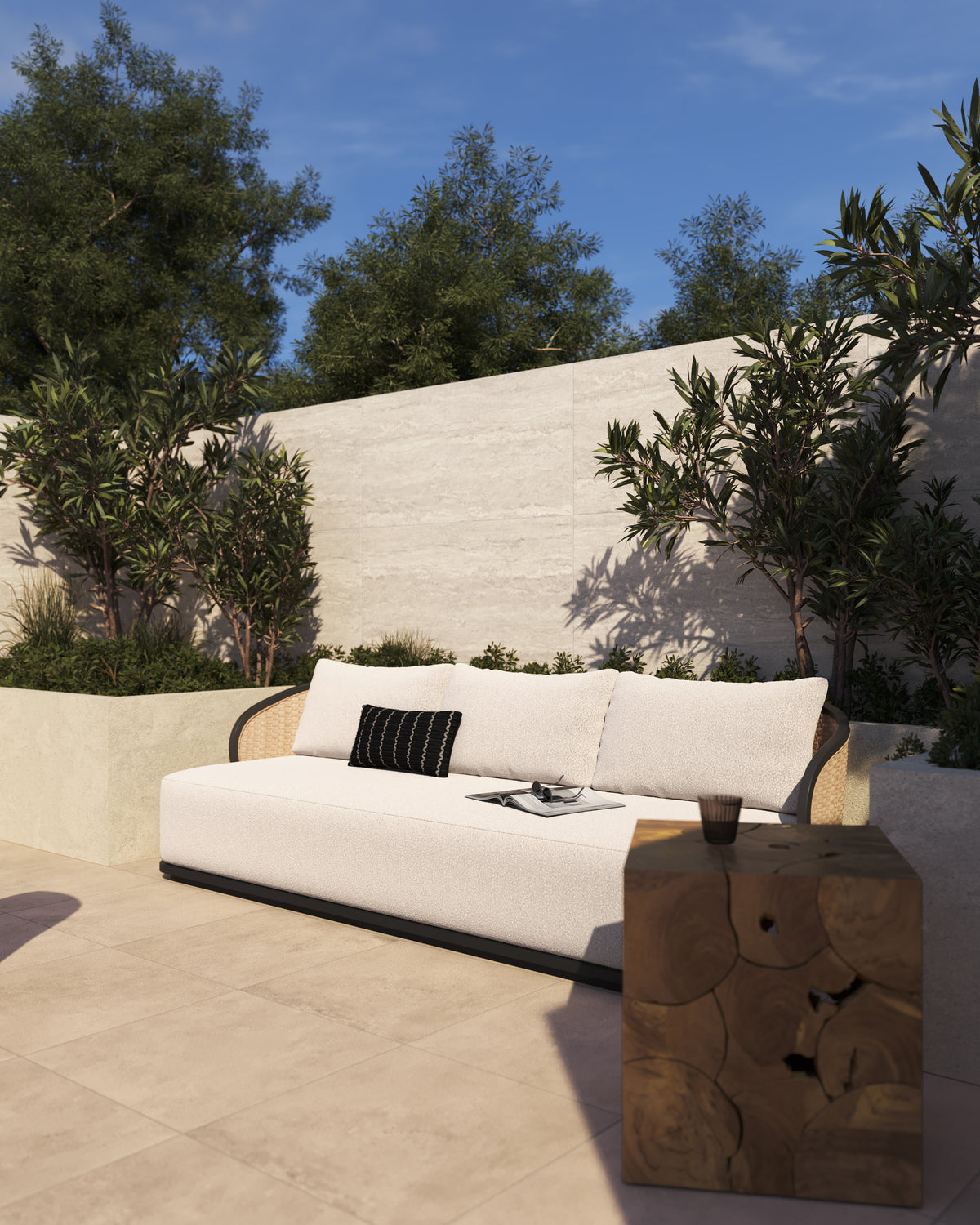 The warm brown teak root mosaic cube of our Cielo Outdoor End Table sits beside a white boucle outdoor sofa on a sun-drenched beige stone patio with green olive trees and a cream wall.