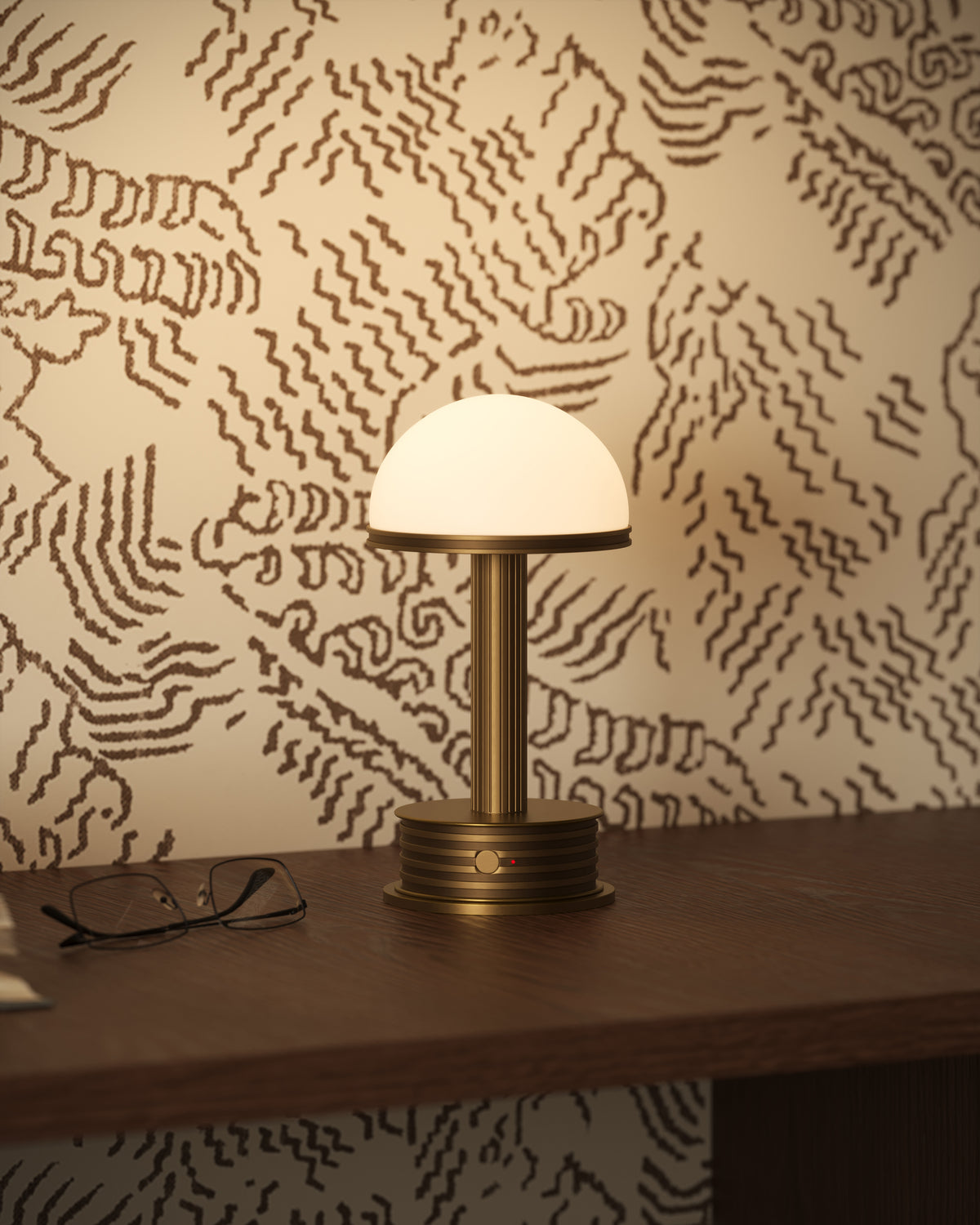 Polished brass portable table lamp with glowing semi-spherical glass shade on a wood desk against abstract patterned wallpaper.