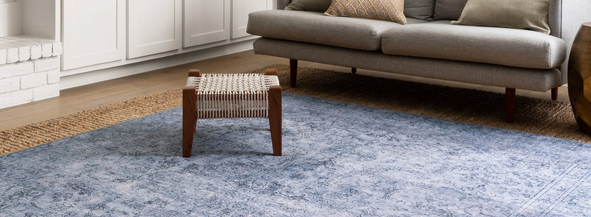 Elegant living space with a cool toned rug featuring intricate patterns, paired with a woven ottoman and a modern gray sofa.