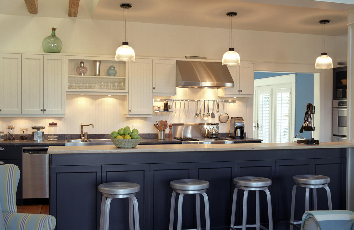 Modern kitchen with three pendant lights over a navy island, stainless steel appliances, and white cabinetry with under-cabinet lighting.