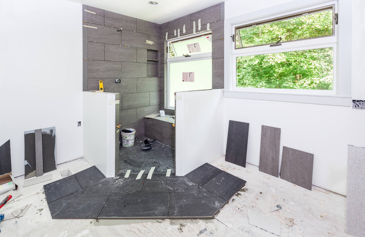 Bathroom under renovation with gray wall tiles, partially installed shower area, loose floor tiles, and tools during tiling process.