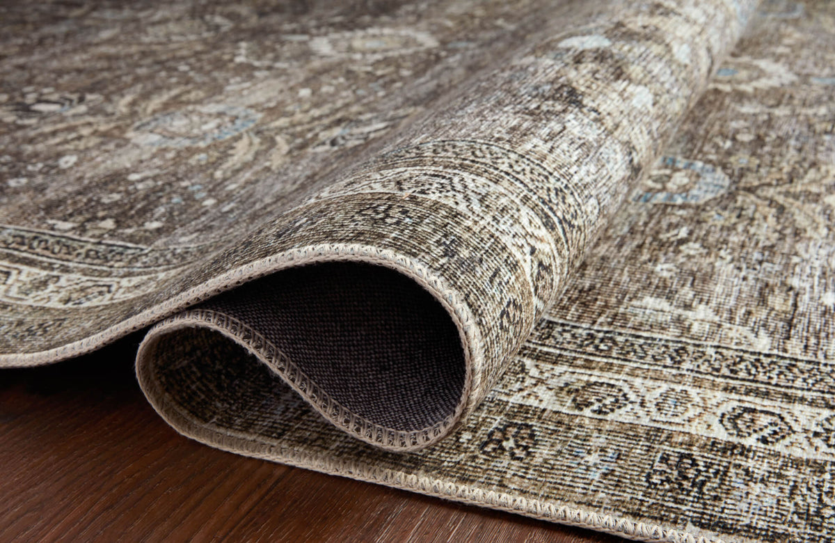Close-up of a rolled traditional-style rug showing a distressed beige and brown pattern, bound edge, woven backing, and low-profile texture on a wood floor.