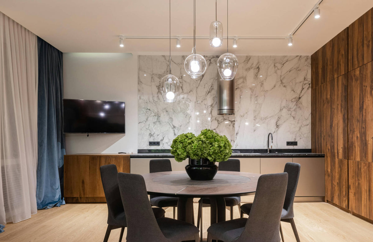 Contemporary open-plan kitchen and dining area featuring clustered glass kitchen pendant lighting above a round dining table, complemented by a marble backsplash, warm wood cabinetry, and sleek modern finishes.