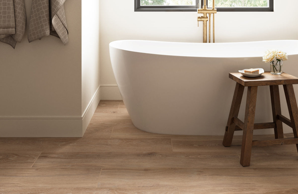 A serene bathroom featuring warm wood look porcelain tile flooring, a sleek white freestanding tub, and elegant brass fixtures.