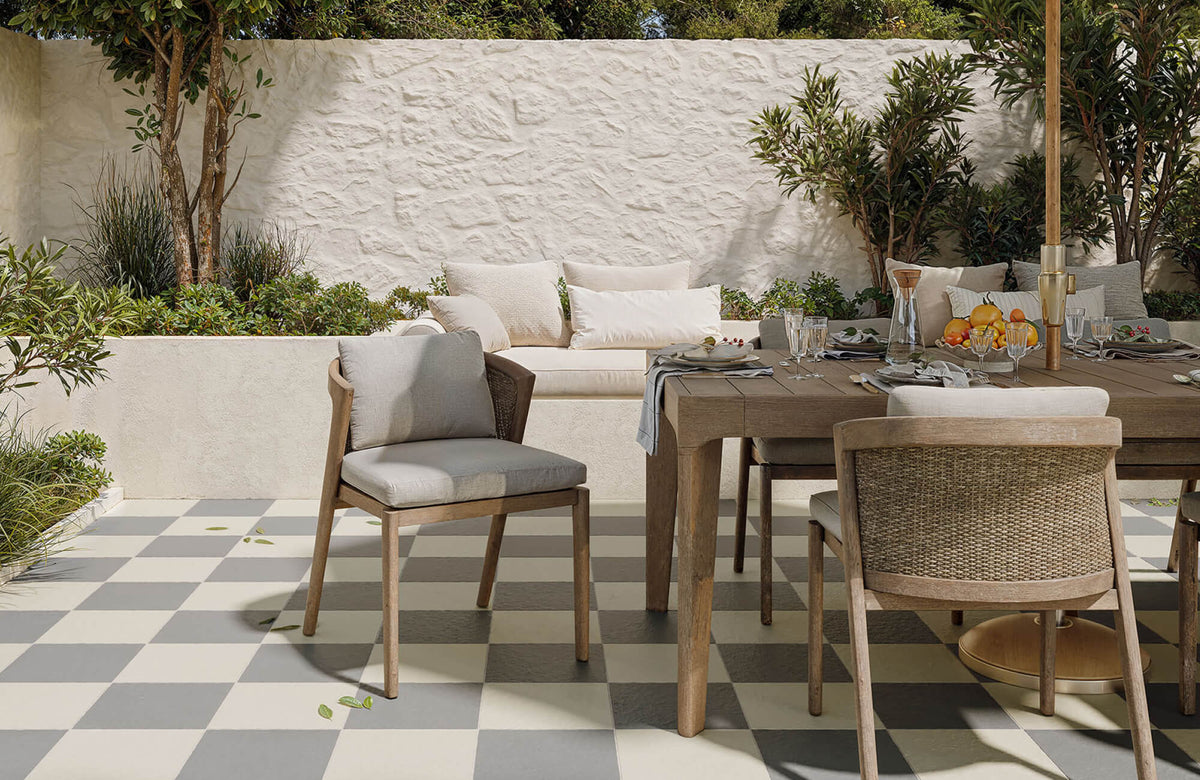 Outdoor dining area with cushioned wood armchairs around a rectangular table on a checkered patio, surrounded by greenery and a white textured garden wall.