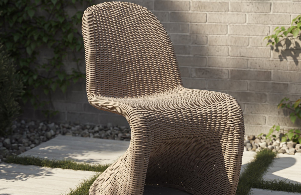 Modern outdoor chair made of tan wicker with a sculpted shape, placed against a neutral brick wall and garden plants.