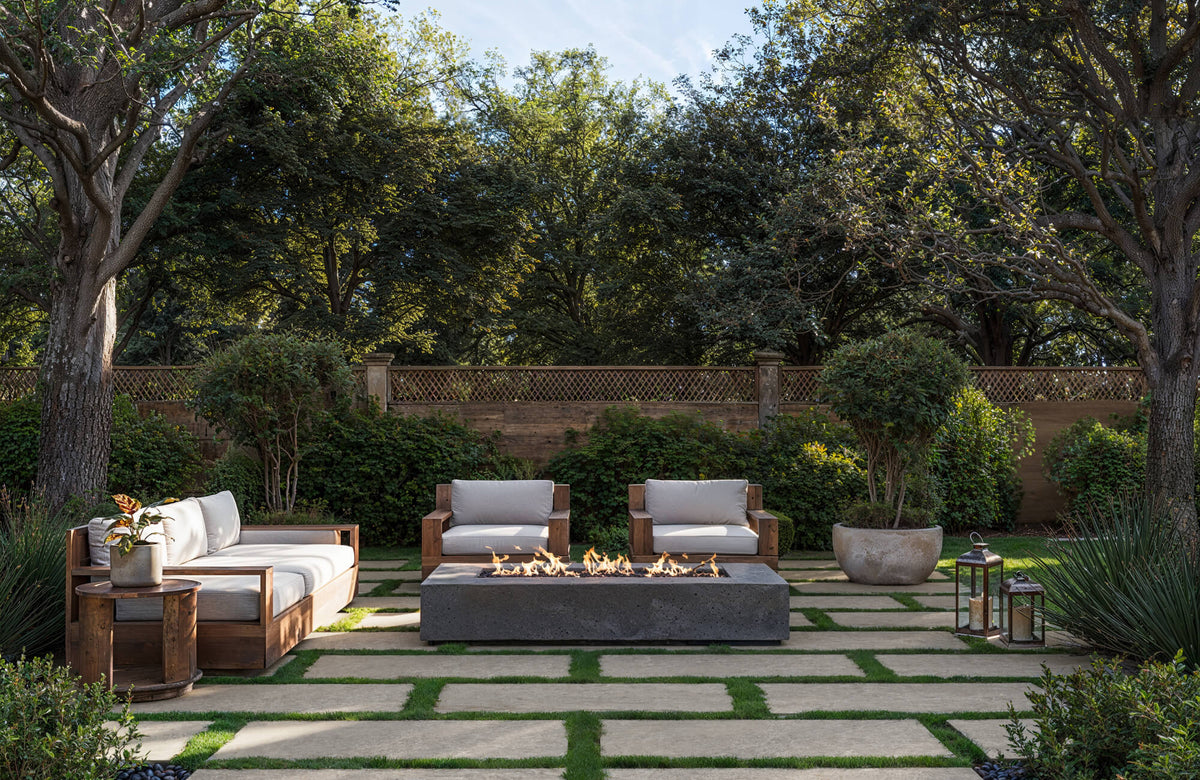 Outdoor seating arranged around a rectangular fire pit with cushioned wood-framed sofas and lounge chairs, surrounded by trees, potted greenery, and stone pavers.