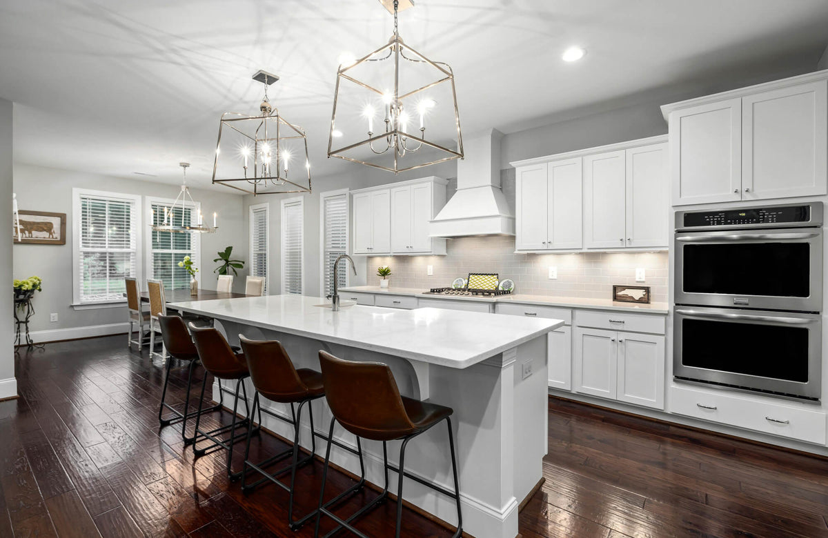 Bright modern kitchen with a large white island, backless bar stools, and geometric pendant lights, surrounded by shaker cabinets and warm wood flooring.