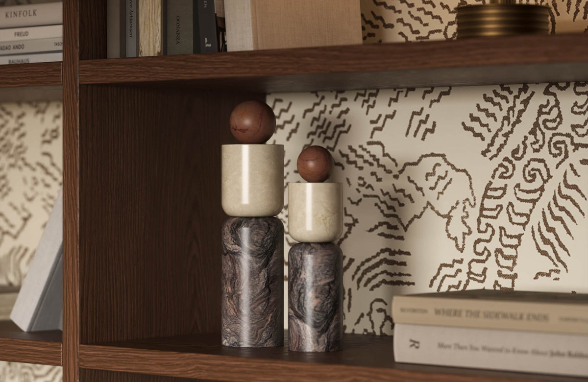 Two stacked marble cylinder sculptures with brown balls on top displayed on a dark wood bookshelf with cream patterned wallpaper and books.