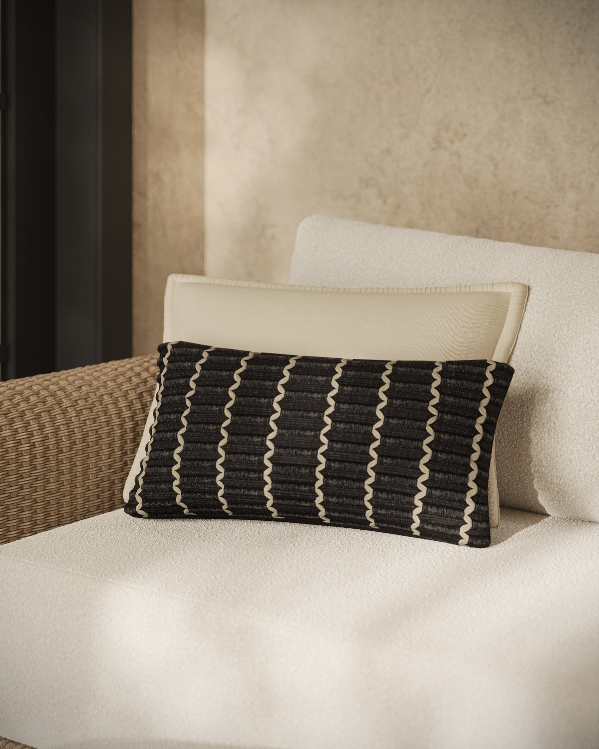 This black accent pillow features vertical ribbed panels with light stitched detailing, layered in front of an off-white pillow on a neutral, textured lounge chair.