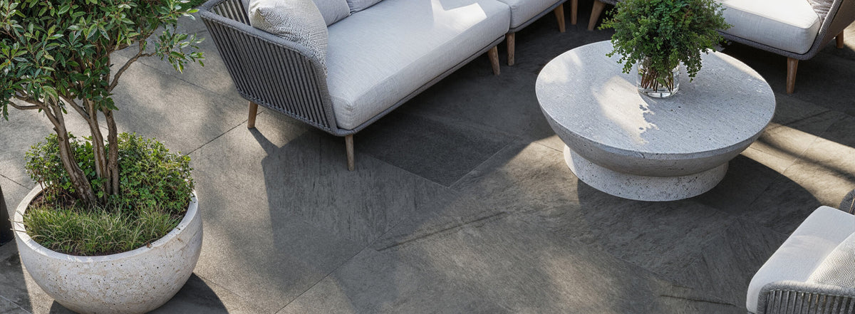 Elegant outdoor seating area with grey stone-look tiles, featuring cozy sofas, lush potted plants, and a round stone table