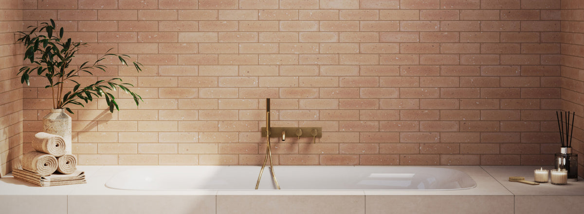 Serene bathroom setting with warm terracotta-toned brick look tiles lining the wall behind a minimalist white bathtub, complemented by gold fixtures and natural décor