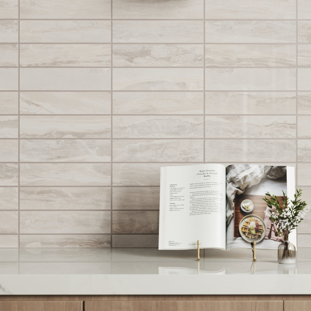 Close-up of Harley Greige 3x12 polished tiles showcasing their elegant, natural veining and soft tones. The polished surface reflects light beautifully, paired with a minimalist countertop featuring an open recipe book and a small vase of fresh flowers, adding charm and sophistication.