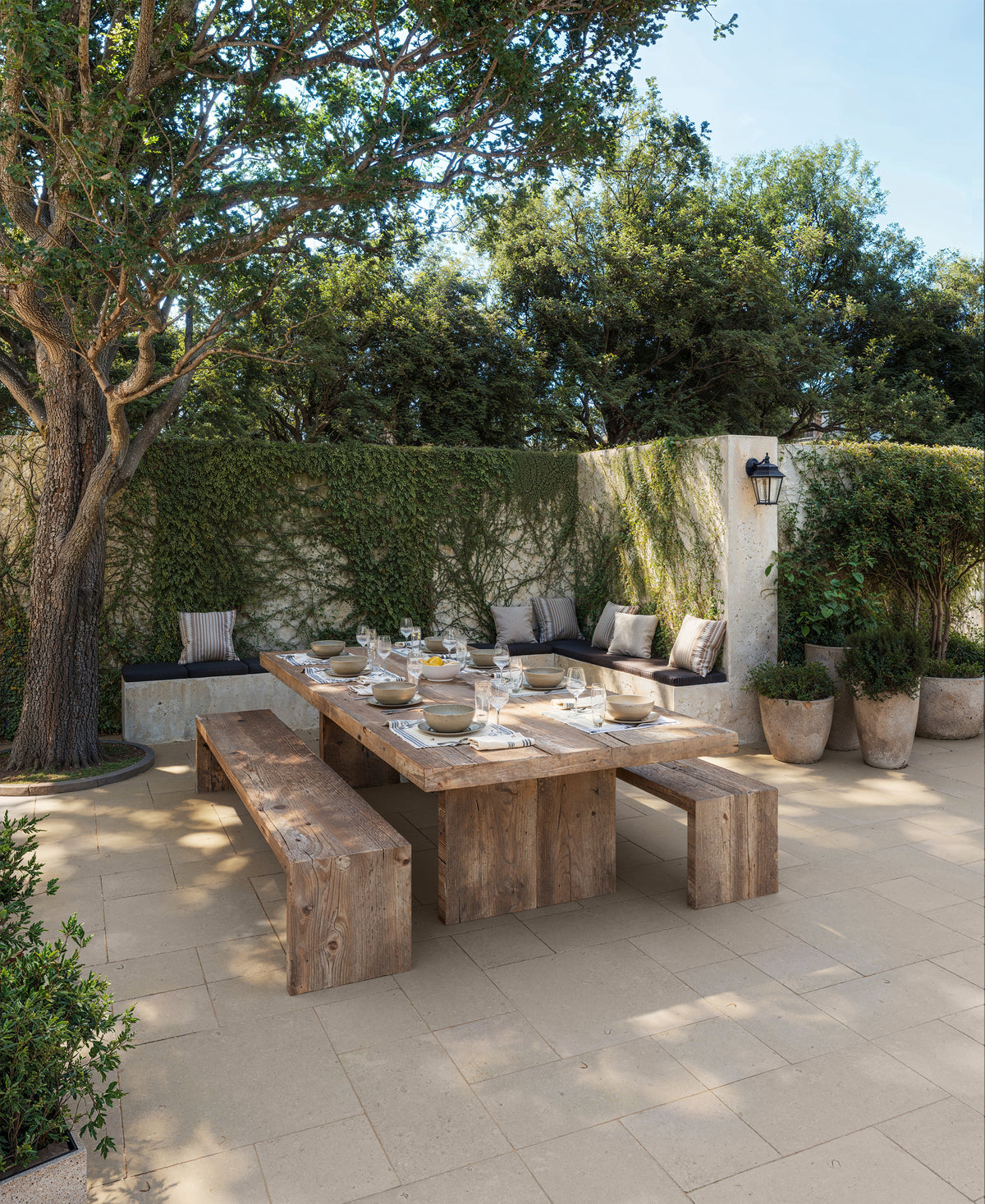 Rustic outdoor dining area featuring 12x24 dune-beige porcelain tiles with a 2cm thickness and matte grip finish, complemented by a wooden table, benches, and lush greenery.